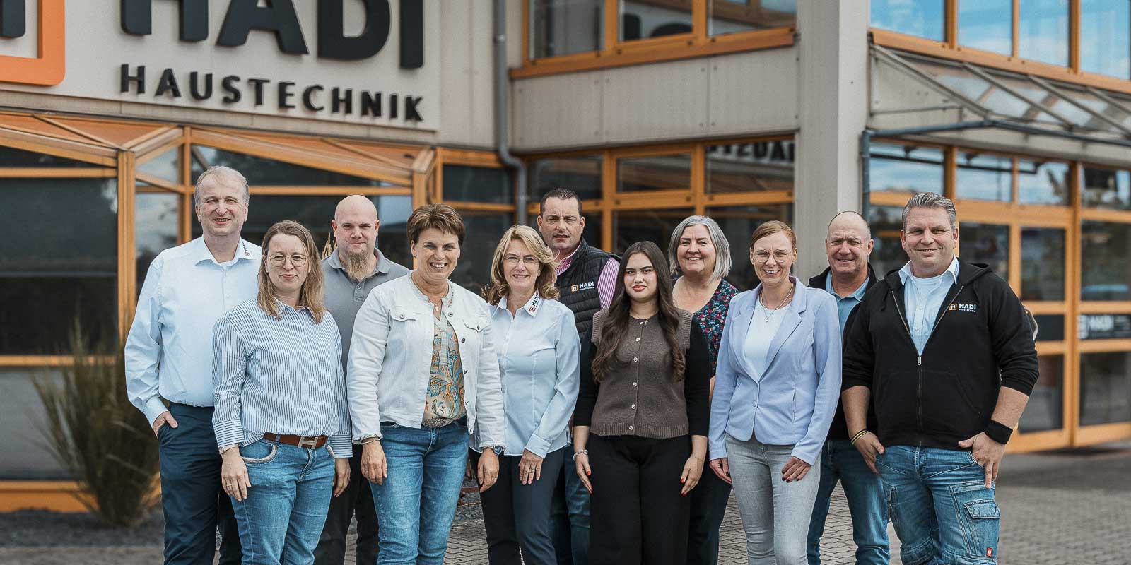 Die Mitarbeiter von HADI Haustechnik posieren in formeller Kleidung vor ihrem Firmengebäude, strahlend und freundlich.