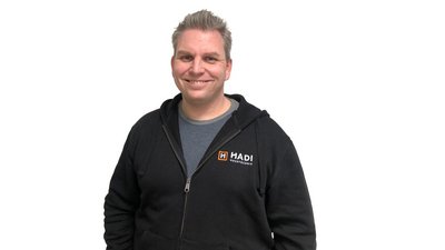 Ein lächelnder Mann trägt einen schwarzen Hoodie mit dem Logo von HADI Maustechik und steht vor einem weißen Hintergrund.