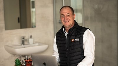 Ein lächelnder Mann in einem modernen Raum sitzt an einem Tisch mit einem Laptop und Schreibutensilien.