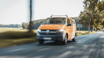 Der VW Bus in leuchtendem Orange bewegt sich dynamisch entlang einer ruhigen Landstraße, die von grünen Feldern gesäumt ist.