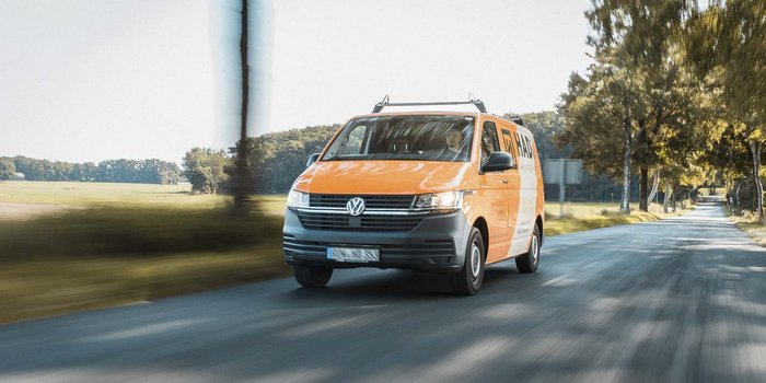 Der VW Bus in leuchtendem Orange bewegt sich dynamisch entlang einer ruhigen Landstraße, die von grünen Feldern gesäumt ist.