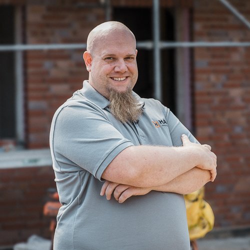 Ein lächelnder Mann mit einem langen Bart steht vor einer Baustelle, umgeben von Ziegelmauern und Baugeräten.