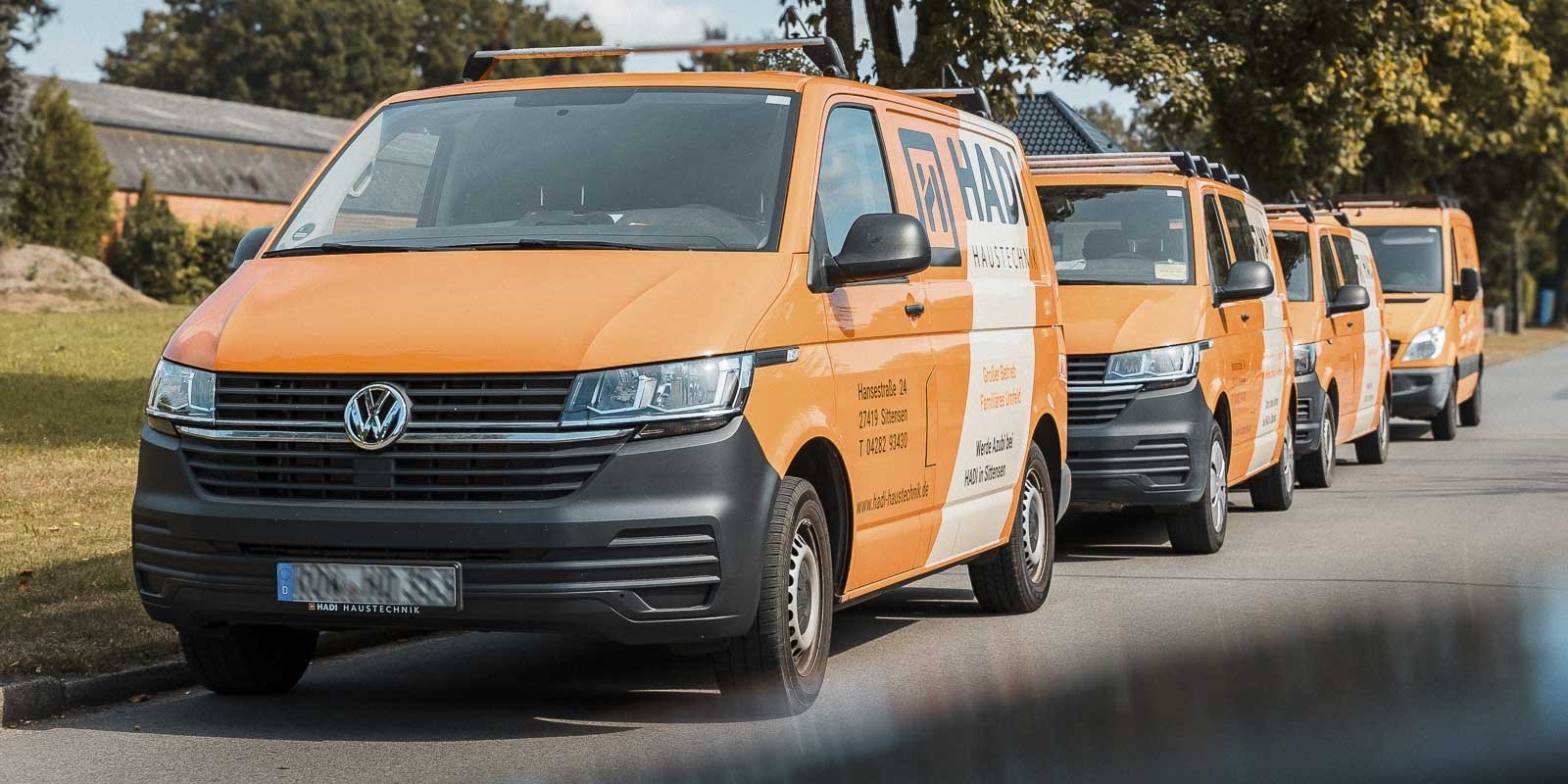 Eine Reihe von orangefarbenen Lieferwagen parkt auf einer Straße, umgeben von Bäumen und einem klaren Himmel.