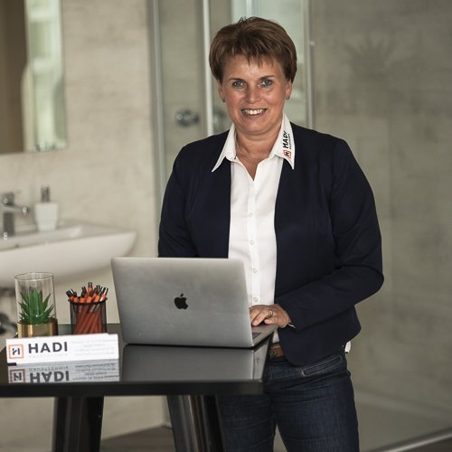 Eine lächelnde Frau steht an einem Tisch mit einem Laptop, umgeben von Büromaterialien und einer modernen Badezimmerumgebung.