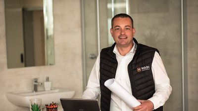 Der Mann trägt eine schwarze Weste mit dem Logo 'HADI' und steht in einem hellen, minimalistischen Raum.