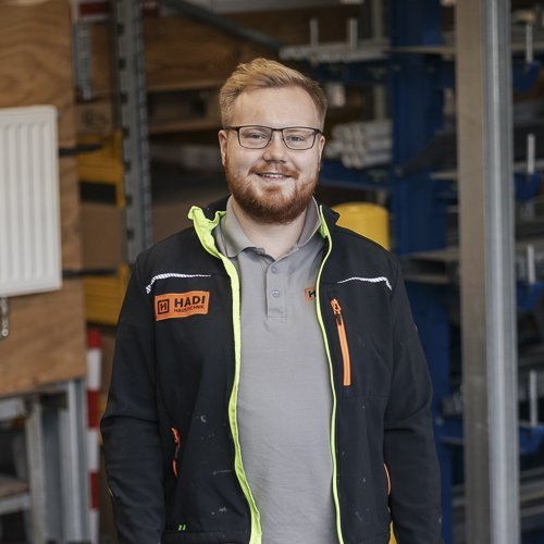 Ein lächelnder Mann mit Brille steht in einer Werkstatt, umgeben von Werkzeugen und Maschinen, in Arbeitskleidung.
