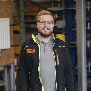 Ein lächelnder Mann mit Brille steht in einer Werkstatt, umgeben von Werkzeugen und Maschinen, in Arbeitskleidung.