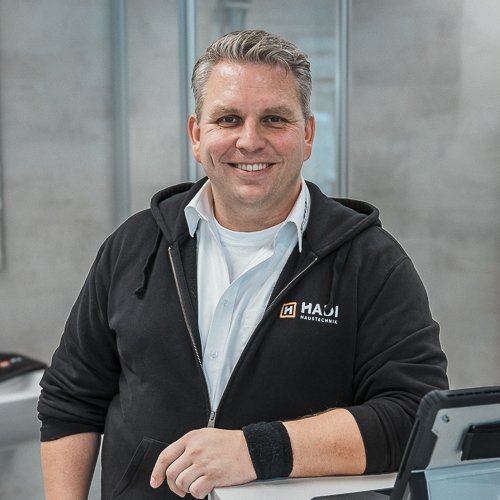 Ein lächelnder Mann in einem schwarzen Hoodie steht an einem Empfangstresen in einem modernen Geschäft.