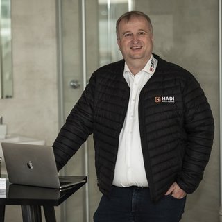 Ein lächelnder Mann in einer schwarzen Jacke steht neben einem Laptop auf einem Tisch in einem modernen Raum.