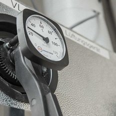 Ein Manometer zeigt eine Temperatur von etwa 60 °C an, montiert an einer grauen Wand mit strukturiertem Muster.