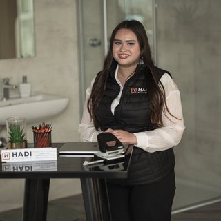 Eine junge Frau mit langen Haaren sitzt an einem Tisch, umgeben von Büromaterialien und einem modernen Badezimmer im Hintergrund.