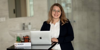 Eine lächelnde Frau mit Brille steht an einem Tisch mit einem Laptop und Büromaterialien in einem modernen Raum.