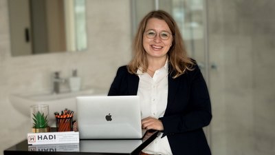 Eine lächelnde Frau mit Brille steht an einem Tisch mit einem Laptop und Büromaterialien in einem modernen Raum.