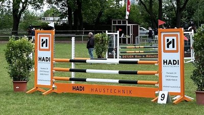 Ein Reitturnier mit einem Sprung, der von orangefarbenen und weißen Stangen umgeben ist, im Hintergrund sind Zuschauer zu sehen.