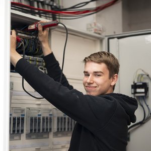 Ein junger Mann arbeitet konzentriert an einem elektrischen Schaltschrank und lächelt dabei freundlich in die Kamera.