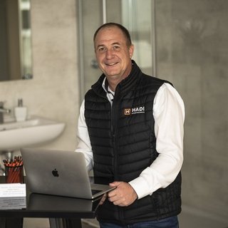 Ein lächelnder Mann in einer schwarzen Weste steht an einem Tisch mit einem Laptop und einem modernen Badezimmer im Hintergrund.