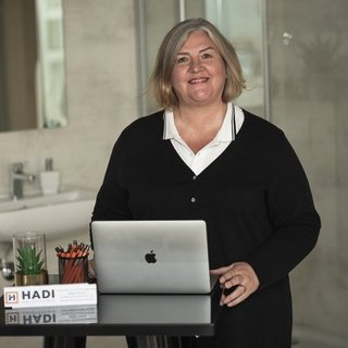 Eine lächelnde Frau mit blonden Haaren steht an einem Tisch mit einem Laptop und Büromaterialien in einem modernen Raum.
