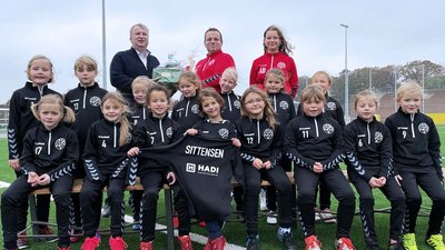 Eine Gruppe von Mädchen in schwarzen Sportanzügen posiert fröhlich auf einer Bank auf einem Fußballplatz.