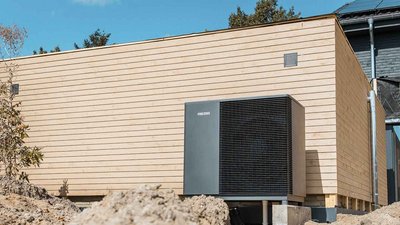Ein modernes Gebäude mit Holzverkleidung, neben dem eine große Wärmepumpe auf einem Erdhügel steht, unter blauem Himmel.