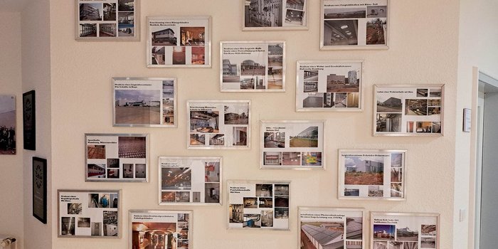 Eine Wand mit mehreren gerahmten Bildern, die verschiedene Bauprojekte und Architekturen zeigen, in einem hellen Raum.