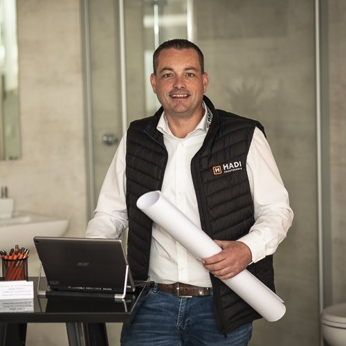 Ein lächelnder Mann in einem schwarzen Weste steht an einem Tisch mit einem Laptop und einem Plan in der Hand.