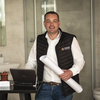 Ein lächelnder Mann in einem schwarzen Weste steht an einem Tisch mit einem Laptop und einem Plan in der Hand.