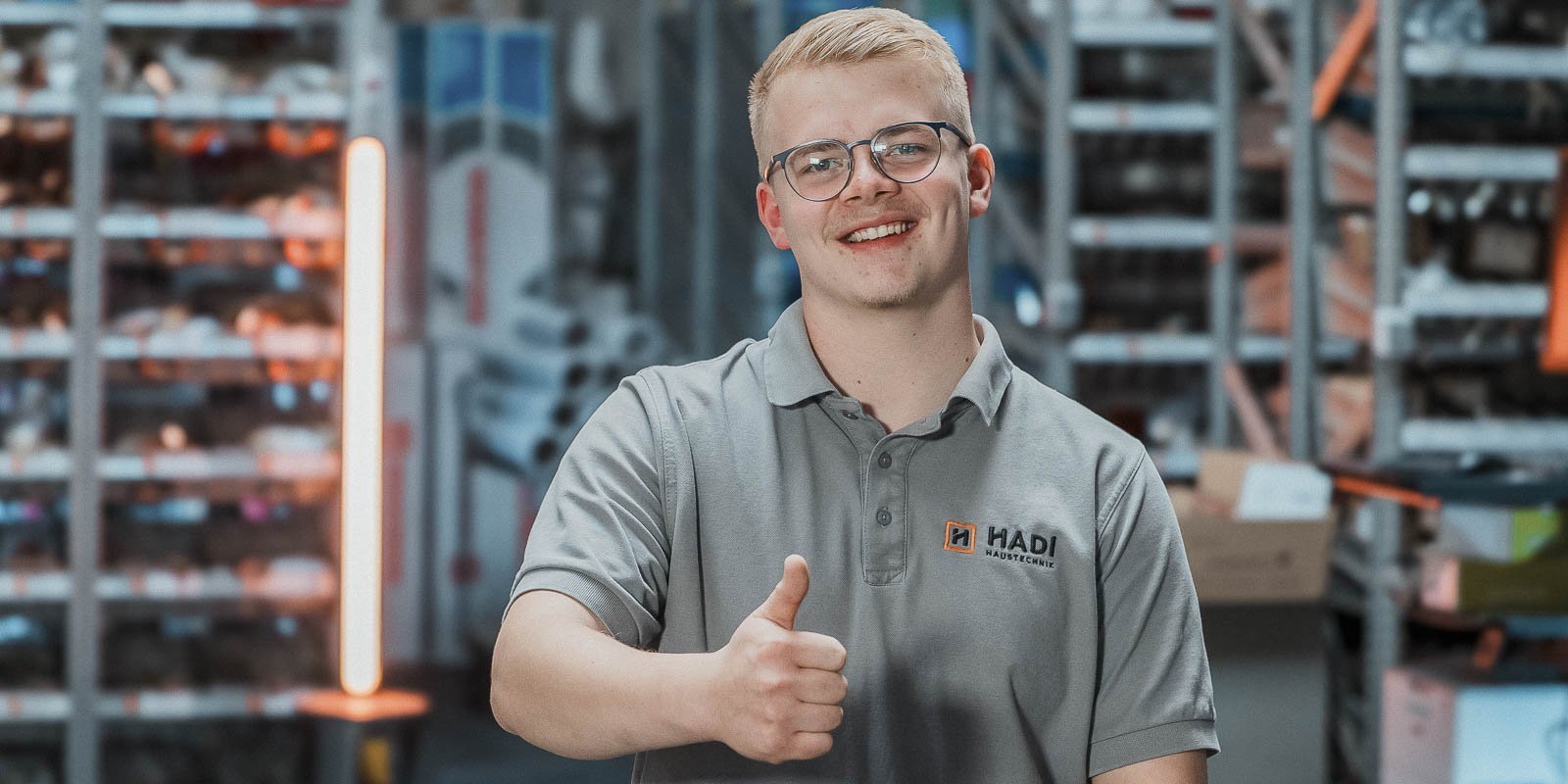 Ein junger Mann mit Brille lächelt und zeigt einen Daumen nach oben, während er in einem Lager mit Regalen steht.