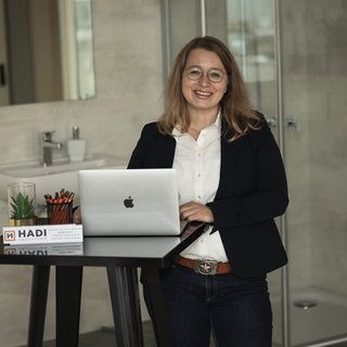 Eine lächelnde Frau steht an einem Tisch mit einem Laptop, umgeben von Büromaterialien und einer Pflanze.