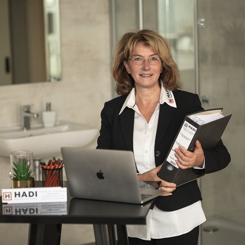 Eine Frau in einem schwarzen Blazer steht an einem Tisch mit einem Laptop und Ordnern, bereit für ein Meeting.