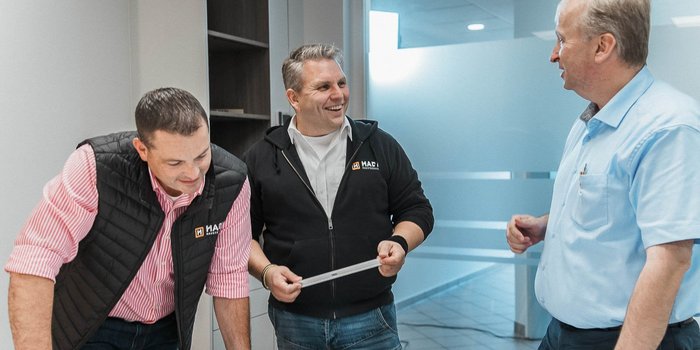 Drei Männer stehen in einem modernen Büro und diskutieren lebhaft, während einer von ihnen ein Maßband hält.