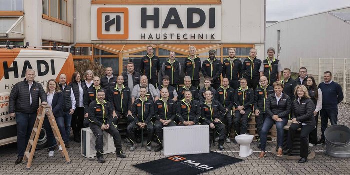 Eine Gruppe von Mitarbeitern der Firma HADI Haustechnik posiert vor ihrem Firmengebäude, umgeben von verschiedenen Materialien.